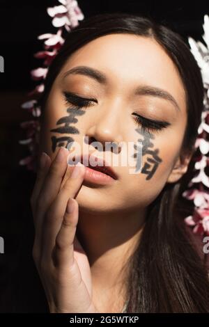 Femme japonaise avec hiéroglyphes sur le visage debout avec les yeux fermés isolés sur le noir Banque D'Images