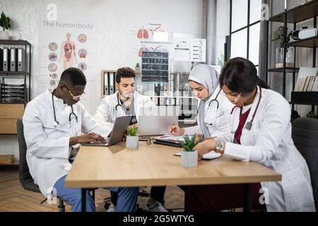 Quatre médecins multiethniques se sont concentrés en conférence à la salle de conférence de l'hôpital. Les travailleurs de la santé, hommes et femmes, utilisent des ordinateurs portables modernes sur des tablettes pendant la réunion. Banque D'Images