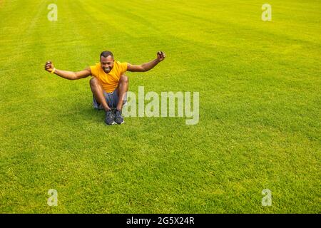 Sorti jeune athlète afro-américain dans des vêtements de sport jaune vif, assis sur l'herbe, souriant célébrant le succès, la victoire. Un mode de vie sain Banque D'Images