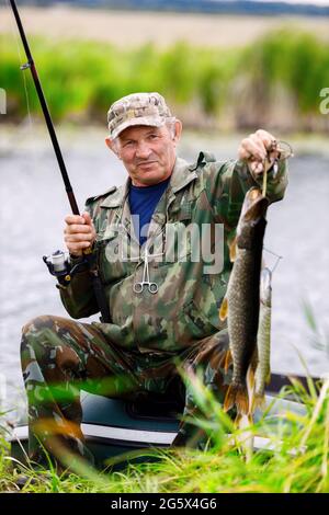 Un pêcheur heureux âgé en camouflage est assis près de la rivière avec une tige de rotation et montre la prise. Ouverture de la saison de pêche. Banque D'Images