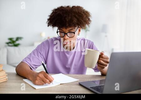 Adolescent noir concentré avec une tasse de café prenant des notes pendant la conférence en ligne sur ordinateur portable à la maison Banque D'Images