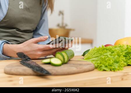 Bonne jeune femme qui boit du jus d'orange, qui navigue sur les réseaux sociaux sur son smartphone tout en cuisinant. Gros plan. Banque D'Images