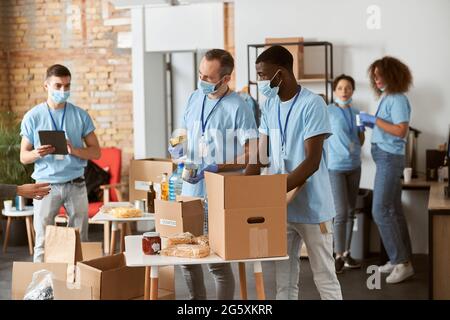 Divers bénévoles en uniforme bleu, masques de protection et gants séparant les produits de donation les uns des autres, en triant les articles dans des boîtes en carton lorsqu'ils sont debout Banque D'Images