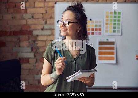 Femme souriante, décoratrice d'intérieur ou architecte en tenue décontractée avec une coiffure désordonnée tenant un stylo et un ordinateur portable, regardant de côté, travaillant sur de nouvelles créations Banque D'Images
