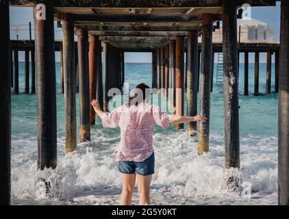 Turquie, Mer méditerranée. Une jeune femme s'est détournée de l'appareil photo, s'éloignant des vagues sous une jetée en bois. Banque D'Images