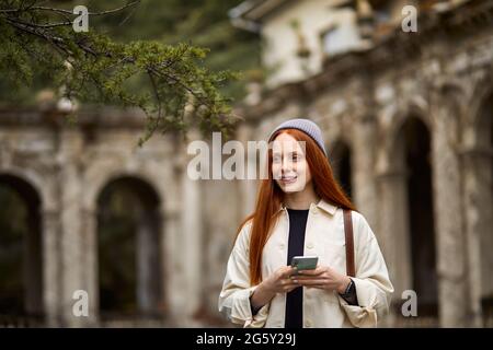Portrait d'une jolie femme se tenant à côté d'un vieux bâtiment en utilisant la technologie tactile au smartphone, en naviguant sur Internet, en regardant à côté, souriant, sp Banque D'Images