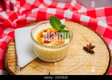 Dessert crème brûlée garni de feuilles de menthe sous forme de ramequin en verre sur une planche en bois et avec une serviette à carreaux rouge-blanc sur un côté Banque D'Images