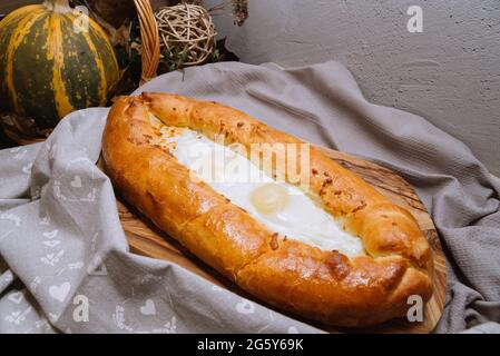 Le khachapuri chaud de style adjarien cuit avec des œufs et du fromage fondu directement du four reposent sur une planche de bois avec une serviette grise à droite Banque D'Images