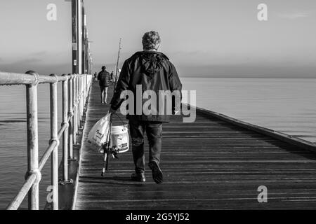 Pêcheur marchant le long d'une jetée en bois sur une mer calme avec des cannes à pêche et un seau à Frankston Victoria Australie Noir et blanc Banque D'Images