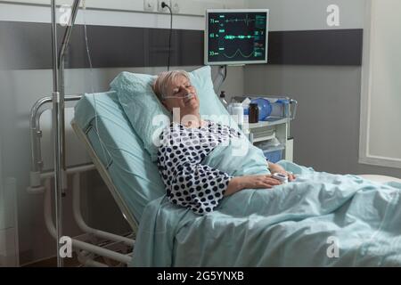 Femme âgée qui se repose dans le lit d'hôpital respire avec l'aide d'un masque à oxygène, en raison d'une maladie pulmonaire. Prendre un médicament intraveineux à partir d'un sac de perfusion iv pendant le traitement de récupération. Banque D'Images