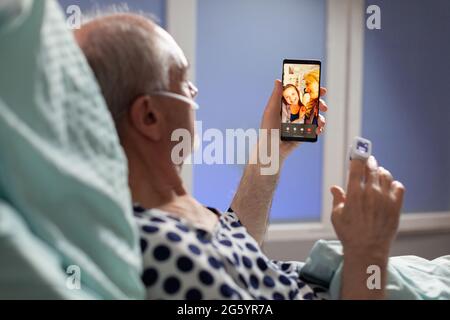 Homme malade senior respirant par tube d'oxygène en disant bonjour à des parents, en posant dans le lit d'hôpital tenant des téléphones, discutant de la récupération. Vieil homme en attente de traitement. Banque D'Images