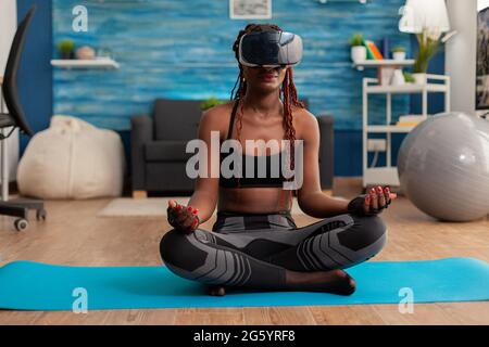Calme tranquille femme noire portant des lunettes de réalité vrituelle assis sur le tapis de yoga, passant du temps sur la spiritualité saine mode de vie. Pratiquer la méditation dans la pose de lotus dans le salon à la maison. Banque D'Images