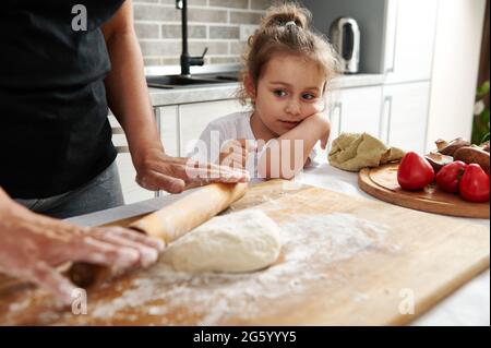 Gros plan d'une petite fille en regardant sa mère rouler la pâte avec un rouleau en bois. Maman et fille cuisent la pizza ensemble. Banque D'Images