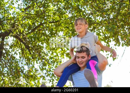 Frère défoqué de la sœur de circonscription à l'arrière. Portrait d'une fille heureuse sur les épaules de l'homme, porcgyback. Petite mouche. Famille jouant à l'extérieur. Flou d'arbre vert Banque D'Images