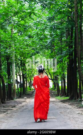 Jeune femme portant une saree rouge marchant sur une route verte en asphalte. La fille marche le long du chemin dans les bois à la lumière. Banque D'Images