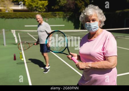 Portrait d'un couple caucasien âgé portant un masque de visage regardant l'appareil photo et souriant sur un court de tennis Banque D'Images