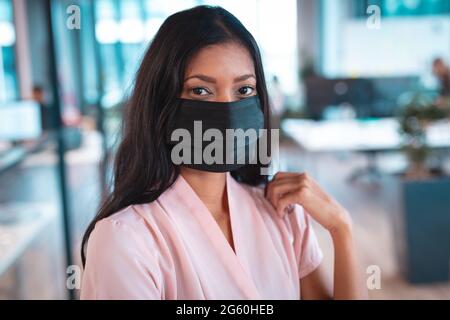 Portrait d'une femme d'affaires de race mixte portant un masque facial avec des collègues en arrière-plan Banque D'Images