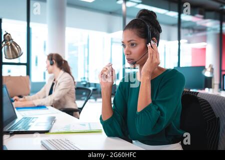 Femme d'affaires de race mixte portant un casque et assis à table parlant, en utilisant un ordinateur Banque D'Images