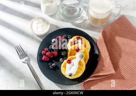 Crêpes au fromage cottage sur une assiette noire avec des bleuets, des fraises et des framboises congelés et une tasse de latte. Petit déjeuner sain. Banque D'Images