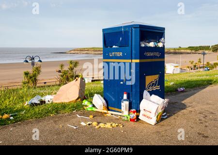 Des déchets et de la nourriture entourent un bac débordant au bord de la mer. Il est tôt le matin, le soleil brille, la plage en arrière-plan est déserte. Banque D'Images