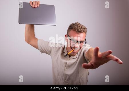 Jeune employé fou avec un ordinateur portable dans la main et un stylo dans la bouche, il porte des lunettes. Homme caucasien avec expression de la folie. Banque D'Images