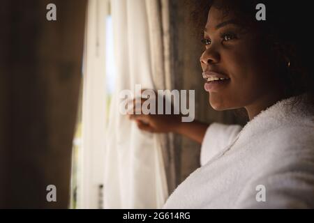 Bonne femme afro-américaine debout à la fenêtre ensoleillée dessinant des rideaux et souriant Banque D'Images