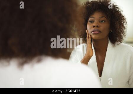 Femme afro-américaine dans la salle de bains portant un peignoir, regardant dans le miroir et hydratant le visage Banque D'Images