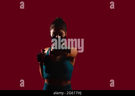 La jeune femme sportive portant des gants de sport est en boxe dans un masque multifonction regardant l'appareil photo sur fond rouge foncé. Banque D'Images