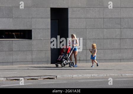 Une famille qui mange de la glace dans la rue. Mère avec ses deux petits. Femme blonde avec glace poussant la poussette avec sa fille à l'intérieur. Banque D'Images