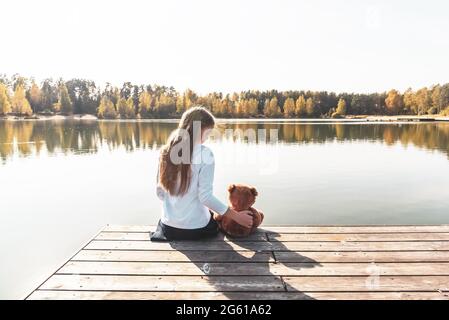 Une fille avec un ours en peluche se trouve sur une jetée en bois près d'une rivière large Banque D'Images