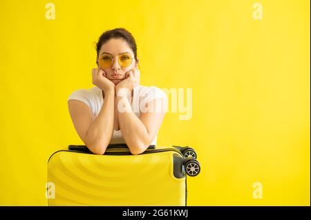 Une femme malheureuse en lunettes de soleil encadre une valise sur fond jaune. Une fille contrariée a manqué son vol dans un avion. Annulation de tous les vols Banque D'Images