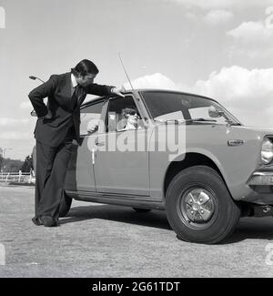 1975, sur une zone pavée hors route, un homme en costume parlant à une femme attirante en lunettes de soleil assis dans son nouveau voiture de Datsun Cherry 100A, Angleterre, Royaume-Uni. Notez le revers large de la veste de costume pour homme et le bas évasé du pantalon et des chaussures plates-formes, des styles de mode distinctifs des années 70. Nissan a commencé à exporter des voitures portant le logo Datsun vers le Royaume-Uni en 1968 et la marque japonaise a connu un grand succès, avec des voitures fiables et abordables, à une époque où l'industrie automobile britannique était en crise, avec des grèves de la main-d'œuvre et une réputation pour faire des voitures d'une mauvaise qualité et fiabilité. Banque D'Images