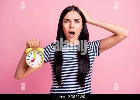 Photo portrait d'une fillette qui se touchante avec une tête tenant une horloge jaune dans une main isolée sur fond rose pastel Banque D'Images