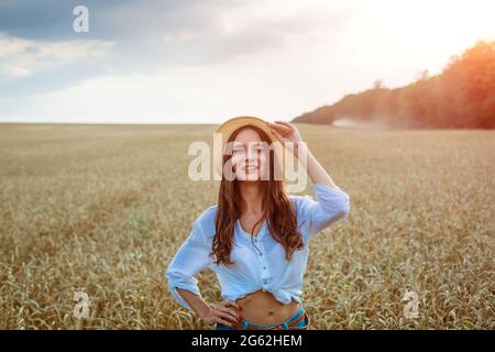 Happy Girl est assis dans un champ de blé en chapeau de paille. Mignon jeune femme d'origine caucasienne en vêtements décontractés aime le blé doré mûr dans un champ. Concept de femme libre Banque D'Images