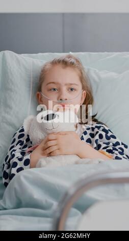 Portrait d'un petit enfant malade solitaire qui se repose dans un lit tenant un ours en peluche dans les mains, regardant la caméra pendant l'examen de récupération à l'hôpital. Enfant hospitalisé en attente de soins de santé Banque D'Images