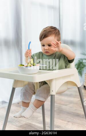 Petit garçon tenant une cuillère près d'un bol de légumes flou sur une chaise haute Banque D'Images