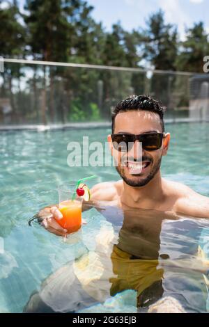 un homme musulman content en lunettes de soleil se détendant dans la piscine avec un cocktail rafraîchissant Banque D'Images