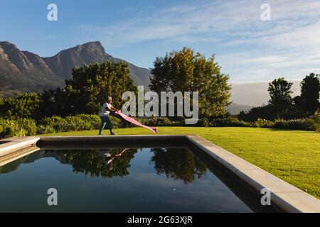 Femme afro-américaine senior mettant le tapis de yoga sur l'herbe au bord de la piscine extérieure dans la campagne Banque D'Images