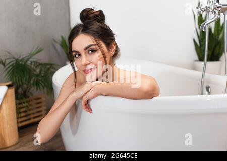 jeune femme gaie avec un petit pain qui regarde l'appareil photo dans la baignoire Banque D'Images