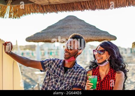 Couple afro-américain prenant le selfie en fête sur la plage - jeunes amis avec masque de visage boire un cocktail en vacances Banque D'Images