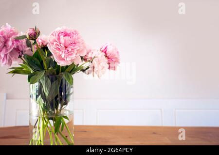 Un bouquet de pivoine rose pastel avec des feuilles vertes dans un vase en verre sur la table en bois près de la texture de mur blanc, décoration lumineuse et confortable pour une maison élégante Banque D'Images