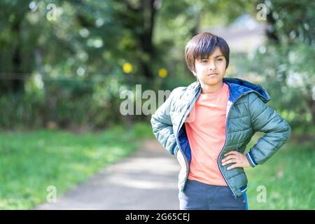 Portrait d'un jeune garçon fatigué à l'extérieur, en lumière naturelle. Enfant d'Asie du Sud portant une veste matelassée avec les mains à la taille en regardant au loin. Banque D'Images