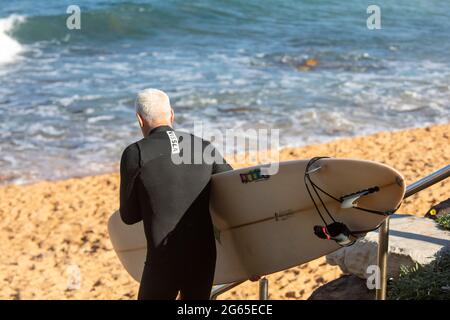 Homme âgé d'un âge moyen aux cheveux gris, portant une planche de surf vers l'océan, Sydney, Australie Banque D'Images