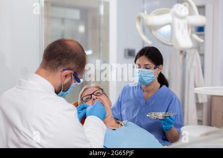 Stomatolog avec assistant de pose de joint dentaire sur les dents de femme âgée. Patient âgé pendant un examen médical avec un dentiste dans un cabinet dentaire équipé d'un équipement orange. Banque D'Images