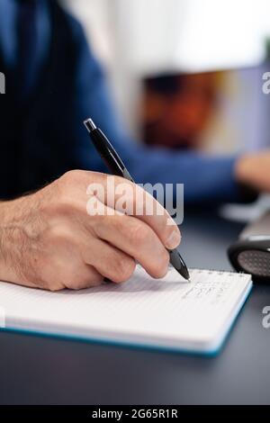 Gros plan sur un homme qui prend des notes sur un ordinateur portable tout en travaillant à la maison. Homme âgé entrepreneur dans le lieu de travail à la maison utilisant un ordinateur portable assis au bureau tandis que la femme lit un livre assis sur le canapé. Banque D'Images