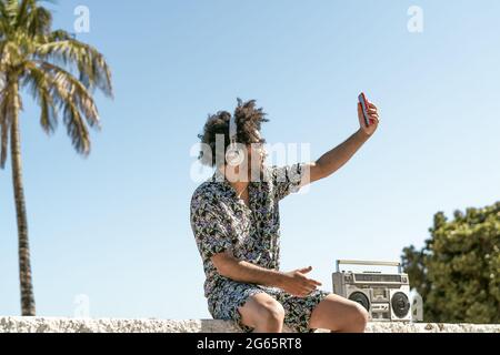 Jeune homme s'amusant à prendre un selfie avec un smartphone mobile tout en écoutant de la musique avec un casque et une boombox vintage pendant les vacances d'été Banque D'Images