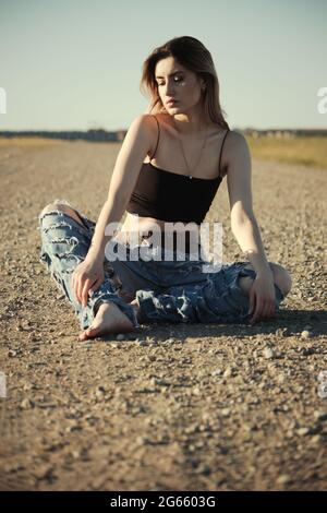 grave triste fille de tramp en jeans déchirés est assis sur une route déserte Banque D'Images