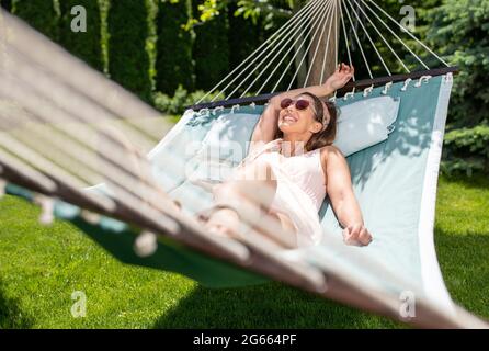 Jeune femme couché dans un hamac se reposant à l'extérieur dans le jardin. Belle fille souriante se relaxant en vacances. Banque D'Images