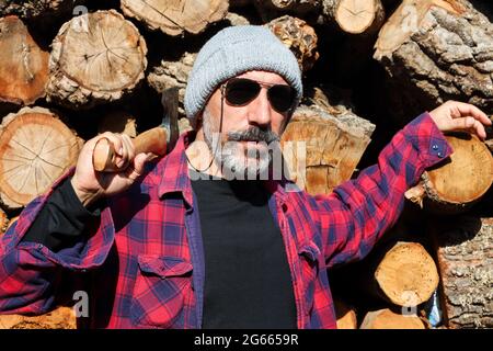 Portrait de magnifique brunette mâle bûcheron avec lunettes de soleil et barbe tenant hache, bois coupé en arrière-plan. Banque D'Images