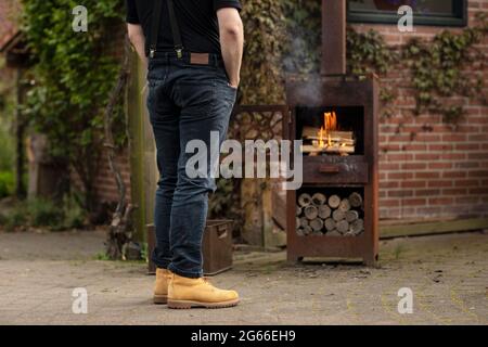 Un homme faisant un feu dans un poêle à bois rouillé et regardant le feu tout en se réchauffant et en appréciant la vie simple dehors dans son jardin Banque D'Images